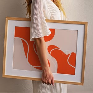 May include: A framed art print with an abstract design. The artwork features orange and white shapes on a light pink background, set within a wooden frame and a white mount.