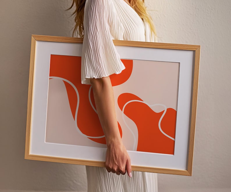 May include: A framed art print with an abstract design. The artwork features orange and white shapes on a light pink background, set within a wooden frame and a white mount.