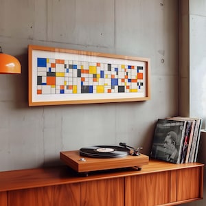 May include: A rectangular framed artwork with a geometric design in primary colours hangs above a wooden record player and cabinet. A stack of vinyl records sits to the right. An orange pendant lamp is visible.