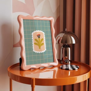 Puede incluir: Un marco de fotos rosa con bordes ondulados que muestra una impresi&oacute;n floral sobre un fondo de cuadr&iacute;cula. Est&aacute; sobre una mesa auxiliar naranja, junto a una l&aacute;mpara plateada y un posavasos a cuadros blanco y negro. Ambiente c&aacute;lido.