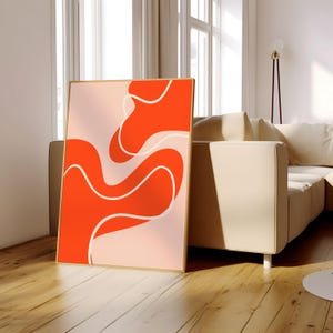 May include: A framed art print with an abstract design in orange and light pink. The artwork features flowing, organic shapes with white outlines, creating a modern visual. The frame is a warm, natural wood tone. The print is leaning against a sofa.