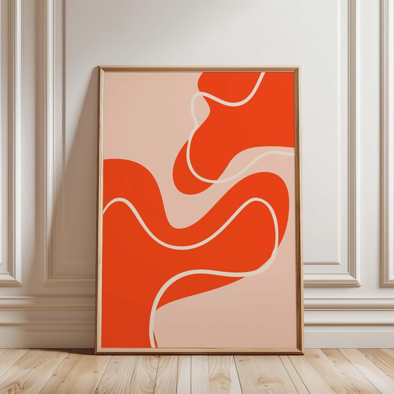 May include: Abstract art print with a bold orange design and white accents set against a pale pink backdrop. The artwork is framed in a natural wood tone, offering a modern, minimalist look. The print is displayed against a white wall and wooden floor.