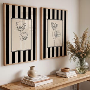 Könnte beinhalten: Zwei gerahmte Wandkunstwerke mit einem schwarz-weiß gestreiften Hintergrund. Jeder Rahmen zeigt ein beiges Rechteck mit einer schwarzen Strichzeichnung von Blumen. Die Rahmen sind aus hellem Holz.