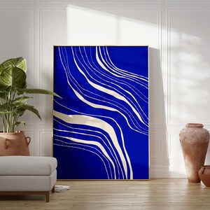 May include: Large framed abstract art print with a vibrant blue background and flowing white lines. The artwork is displayed in a bright room with a potted plant, a grey ottoman, and terracotta vases.