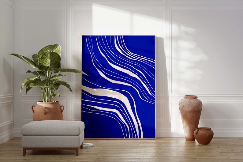 May include: Large framed abstract art print with a vibrant blue background and flowing white lines. The artwork is displayed in a bright room with a potted plant, a grey ottoman, and terracotta vases.