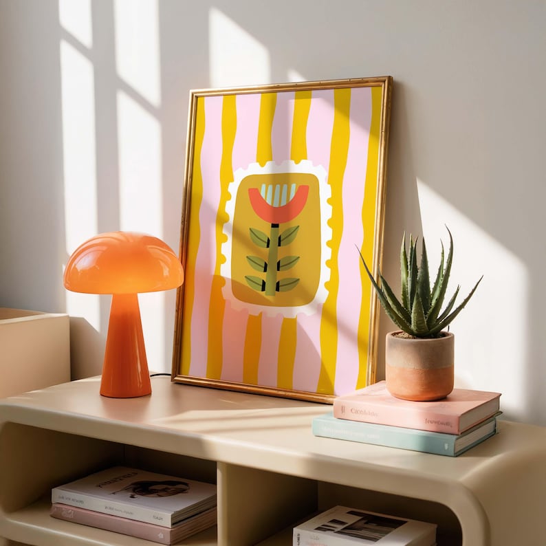 Puede incluir: Una impresi&oacute;n art&iacute;stica enmarcada con un marco dorado que presenta un dise&ntilde;o floral colorido sobre un fondo a rayas. Una l&aacute;mpara naranja con forma de hongo y una suculenta en maceta se encuentran sobre una mesa beige, junto a libros apilados. La obra de arte a&ntilde;ade un toque de color a la habitaci&oacute;n.