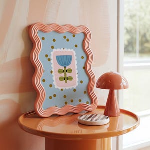 Puede incluir: Una composici&oacute;n decorativa con una impresi&oacute;n floral enmarcada, una l&aacute;mpara con forma de hongo y un posavasos a cuadros sobre una mesa redonda. El marco tiene un borde ondulado y un fondo azul claro con lunares dorados.