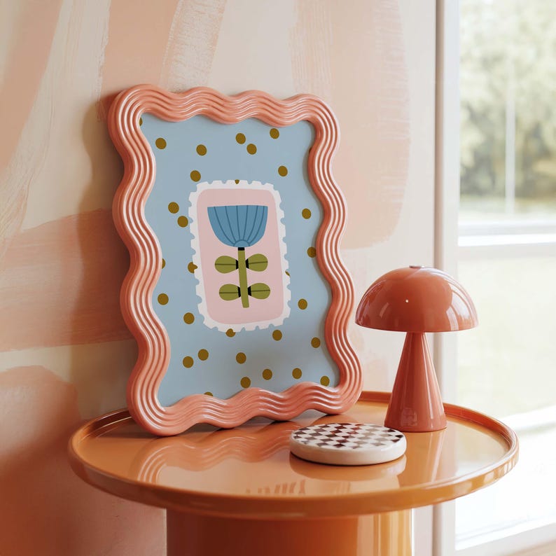 Puede incluir: Una composici&oacute;n decorativa con una impresi&oacute;n floral enmarcada, una l&aacute;mpara con forma de hongo y un posavasos a cuadros sobre una mesa redonda. El marco tiene un borde ondulado y un fondo azul claro con lunares dorados.