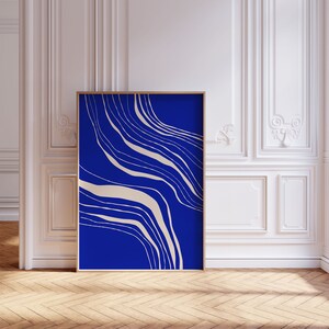 May include: An art print in a wooden frame, showcasing an abstract design. The artwork has a deep blue backdrop with flowing, beige lines. The print is displayed in a room with white walls and a wooden floor.