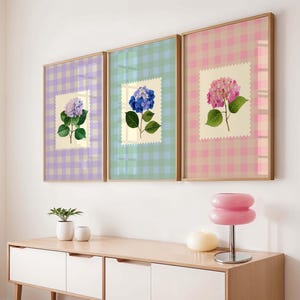Op de afbeelding: Drie ingelijste bloemenprints met hortensiabloemen. Elke print heeft een ruitjesachtergrond in pastelkleuren: lavendel, mintgroen en roze. De hortensiabloemen zijn in tinten paars, blauw en roze. De frames zijn licht van kleur.