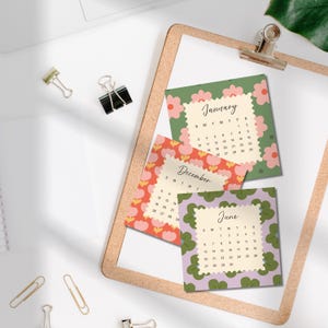May include: Three square calendar cards featuring floral patterns in green, red, and purple. Each card displays a specific month, January, December, and June, with a central cream-colored grid for dates. The cards have scalloped edges and are arranged on a clipboard.