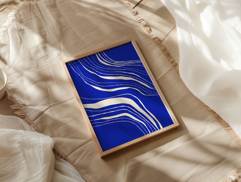 May include: A framed art print with a striking blue backdrop and cream-coloured, flowing lines. The artwork is presented in a light-toned wooden frame, offering a modern, minimalist look. The print rests on a beige fabric surface.
