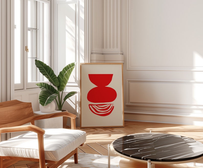 May include: A red abstract art print in a wooden frame hangs on a white wall above a wooden chair and a black marble coffee table. The print features a stack of red geometric shapes.