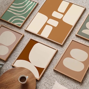 Puede incluir: Una colección de láminas artísticas abstractas enmarcadas en varios colores, incluyendo verde, marrón y rosa, con formas geométricas beige. Las láminas están expuestas sobre una alfombra clara, con una mesa de madera y una taza de café en primer plano.