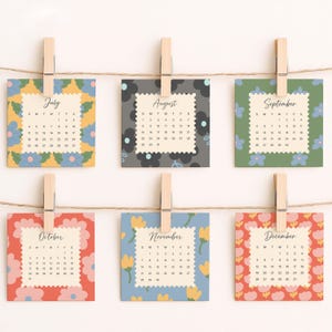 May include: Six square calendar cards for July through December, each featuring a unique floral border pattern and a scalloped-edge center with month names and dates. The cards are suspended by wooden clothespins on a thin brown twine string.