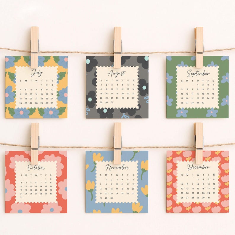 May include: Six square calendar cards for July through December, each featuring a unique floral border pattern and a scalloped-edge center with month names and dates. The cards are suspended by wooden clothespins on a thin brown twine string.