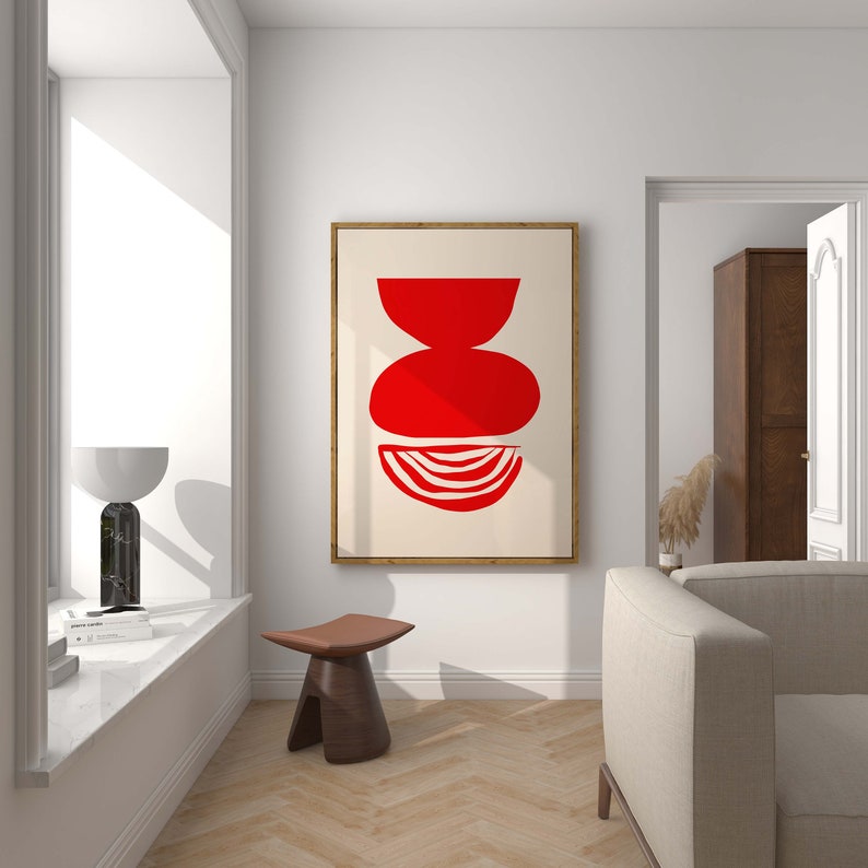 May include: A framed abstract art print with a red geometric design on a cream background. The print is hanging on a white wall in a living room setting.