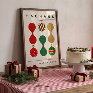 May include: A framed poster with the text BAUHAUS and JULI-SEPT 1923, featuring a graphic design of red, green, and gold abstract shapes and Christmas ornaments. The poster is displayed alongside a frosted cake and wrapped gift boxes.