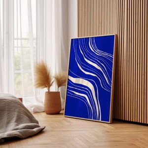 May include: A framed art print with a striking blue backdrop and flowing cream abstract lines. The artwork is displayed in a room with natural light, a woven basket with decorative plants, and wooden paneling.