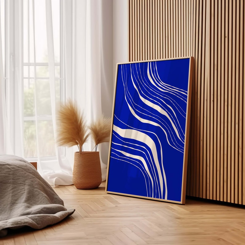 May include: A framed art print with a striking blue backdrop and flowing cream abstract lines. The artwork is displayed in a room with natural light, a woven basket with decorative plants, and wooden paneling.