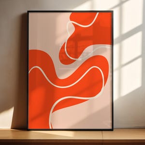 May include: Abstract art print in a black frame, featuring an orange design with white lines on a pale pink background. The artwork is displayed on a wooden surface against a neutral wall. The design has a modern, minimalist aesthetic.