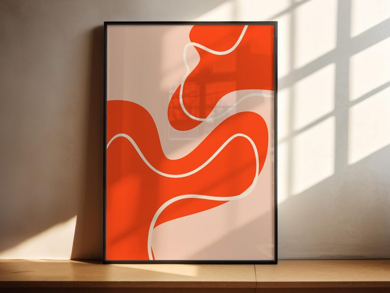 May include: Abstract art print in a black frame, featuring an orange design with white lines on a pale pink background. The artwork is displayed on a wooden surface against a neutral wall. The design has a modern, minimalist aesthetic.