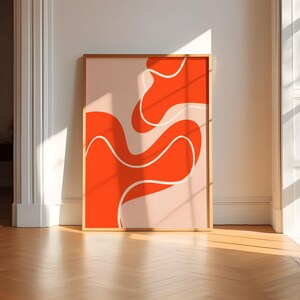 May include: A framed abstract art print with a bold orange design and white and light pink details. The artwork has a contemporary style and is set against a white wall and wooden floor.