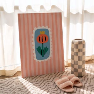 Puede incluir: Una impresi&oacute;n art&iacute;stica decorativa con un fondo a rayas rosas y blancas. La impresi&oacute;n presenta un dise&ntilde;o floral dentro de una forma azul similar a un sello. Tambi&eacute;n se ven un jarr&oacute;n a cuadros y zapatillas rosas.
