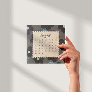 May include: A square calendar page for August featuring a cream-colored center with a scalloped edge, surrounded by a dark gray background with black flower silhouettes and light blue accents. The text August is printed in a script font.