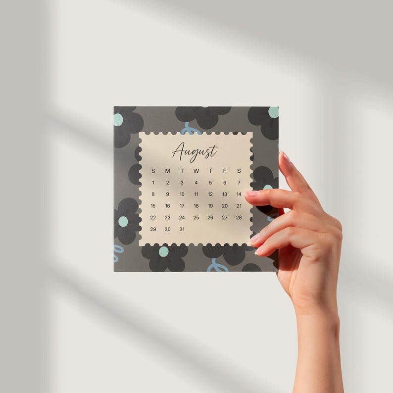May include: A square calendar page for August featuring a cream-colored center with a scalloped edge, surrounded by a dark gray background with black flower silhouettes and light blue accents. The text August is printed in a script font.