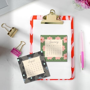 May include: A clipboard with a red and pink heart pattern, a gold clip, and two square calendar pages. One page shows January with pink flowers on a green background, and the other shows August with black flowers on a dark gray background.