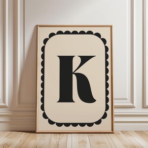 Op de afbeelding: Een ingelijste kunstdruk met de zwarte letter "K" op een crèmekleurige achtergrond. De letter is gecentreerd in een zwarte geschulpte rand, alles in een licht houten frame. Het kunstwerk wordt gepresenteerd tegen een witte muur en een houten vloer.