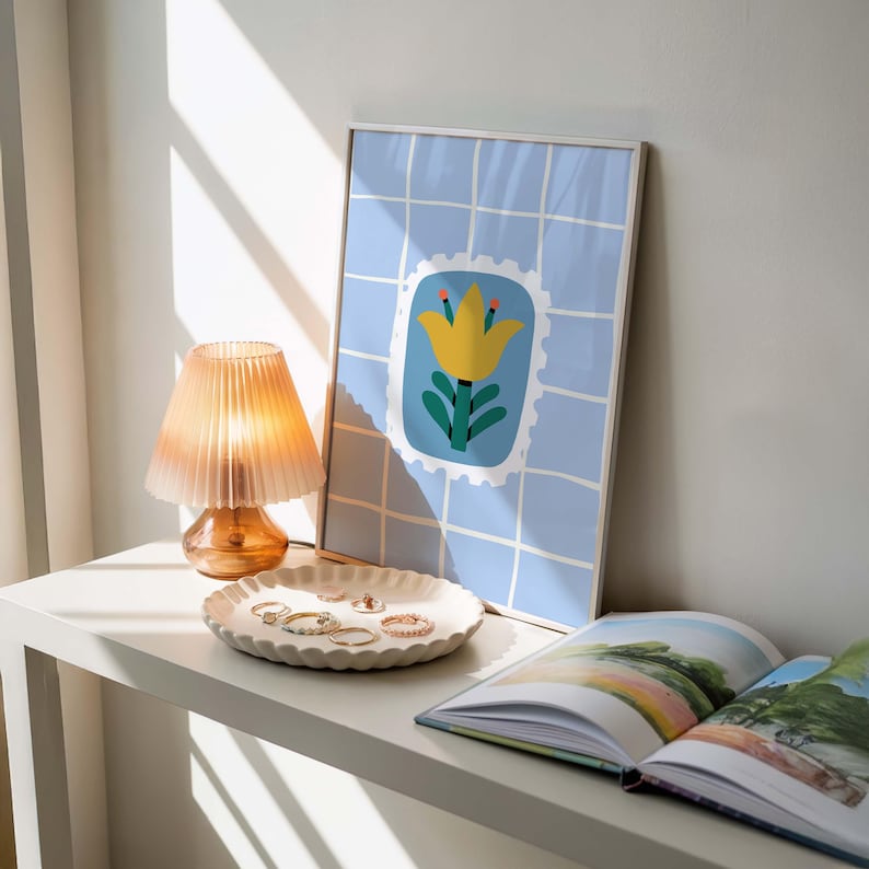 Puede incluir: Una impresi&oacute;n art&iacute;stica enmarcada con un dise&ntilde;o de flor amarilla sobre un fondo de cuadr&iacute;cula azul. Una peque&ntilde;a l&aacute;mpara de color &aacute;mbar con una pantalla plisada se encuentra junto a un plato blanco decorativo que contiene varios anillos. Tambi&eacute;n hay un libro abierto sobre la mesa.