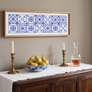 Könnte beinhalten: Ein gerahmtes Kunstwerk mit blau-weißem Fliesenmuster hängt über einem Holzschrank. Eine Schüssel mit Birnen, zwei Messingkerzenhalter mit weißen Kerzen und eine Glaskaraffe mit bernsteinfarbener Flüssigkeit stehen auf dem Schrank.