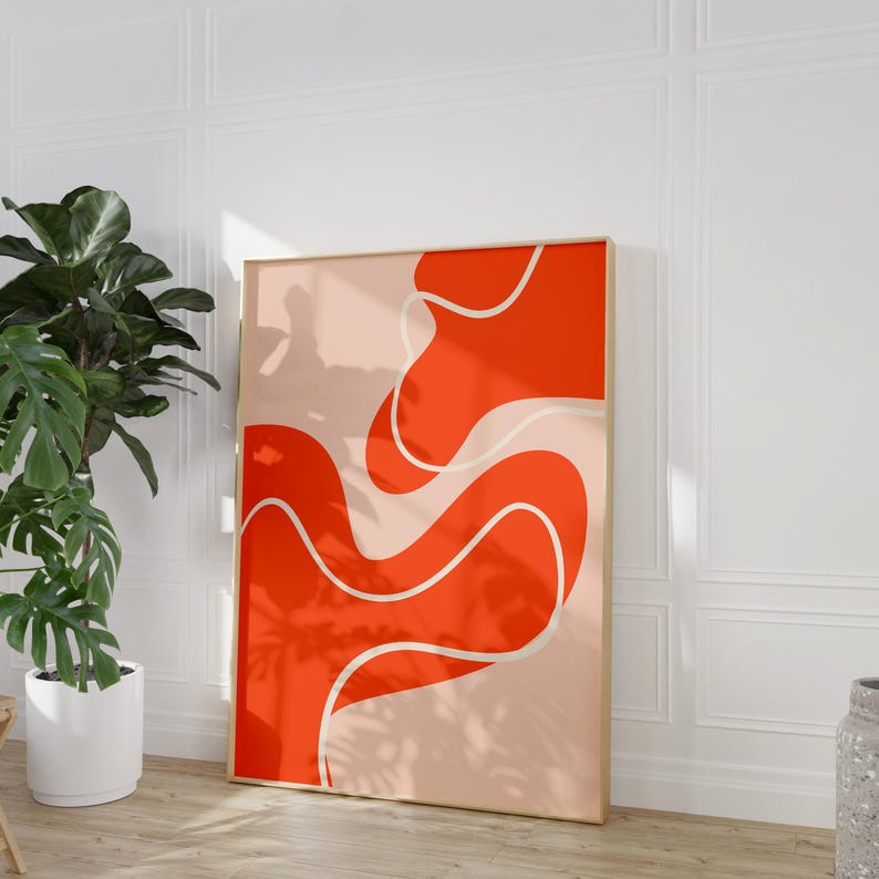 May include: A framed abstract art print with a bold orange design and white outlines on a peach backdrop. The artwork, in a gold frame, leans against a white wall. The overall style is modern and minimalist.