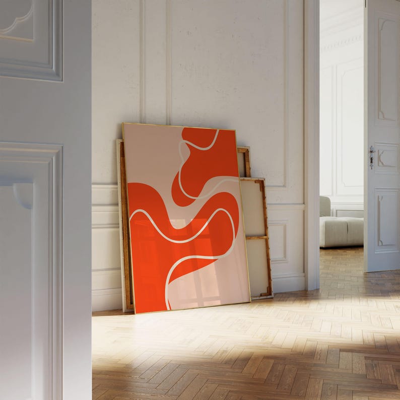 May include: An abstract art print with a bold orange design and white detailing against a pale pink backdrop. The artwork, in a gold frame, leans against a wall. The style is contemporary and minimalist, suitable for modern interiors.