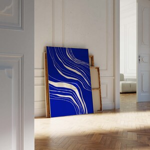 May include: A large framed art print with a striking blue backdrop and flowing white abstract lines. The artwork, with its gold frame, leans against a white wall, creating a modern, minimalist aesthetic. The print is a statement piece.