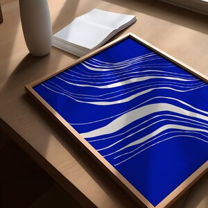 May include: A framed art print with a striking blue backdrop and flowing white lines. The artwork is presented on a wooden surface, with a white vase and a stack of blank pages. The frame is a light brown colour.