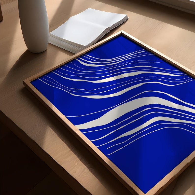 May include: A framed art print with a striking blue backdrop and flowing white lines. The artwork is presented on a wooden surface, with a white vase and a stack of blank pages. The frame is a light brown colour.