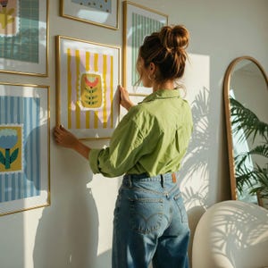 Puede incluir: Una persona cuelga impresiones art&iacute;sticas enmarcadas en una pared blanca. El arte presenta dise&ntilde;os florales y abstractos coloridos en marcos dorados. La persona lleva una camisa verde y vaqueros azules. Un espejo grande y una silla blanca son visibles en el fondo.