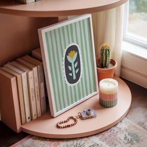 Puede incluir: Una impresi&oacute;n art&iacute;stica enmarcada con un dise&ntilde;o floral, colocada en una estanter&iacute;a rosa. La impresi&oacute;n presenta una flor amarilla y hojas verdes sobre un fondo a rayas verdes y blancas. Un cactus, una vela y objetos decorativos completan la exhibici&oacute;n.