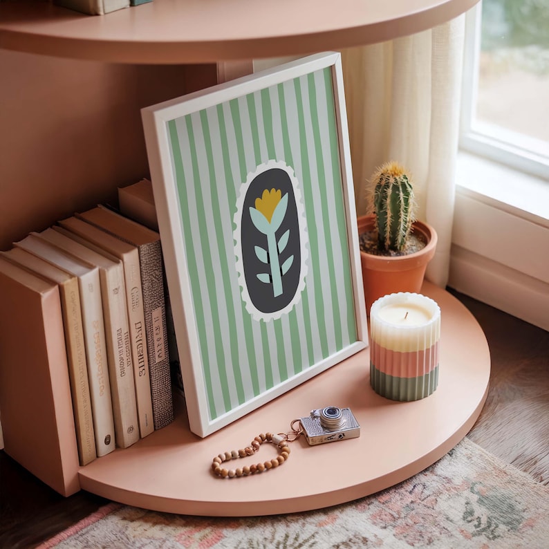 Puede incluir: Una impresi&oacute;n art&iacute;stica enmarcada con un dise&ntilde;o floral, colocada en una estanter&iacute;a rosa. La impresi&oacute;n presenta una flor amarilla y hojas verdes sobre un fondo a rayas verdes y blancas. Un cactus, una vela y objetos decorativos completan la exhibici&oacute;n.