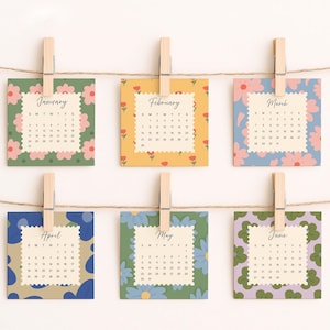 May include: Six square calendar cards for January through June, each featuring a unique floral pattern and scalloped edges. The cards are held by wooden clothespins on a string. Each card displays the month name and a grid of dates.