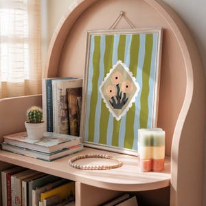Puede incluir: Una obra de arte enmarcada con un fondo a rayas verdes y azules y un dise&ntilde;o floral en forma de diamante. La obra de arte se exhibe en una estanter&iacute;a rosa con libros, un peque&ntilde;o cactus, una pulsera de cuentas y una vela en capas.