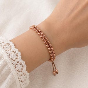 May include: A rose gold-toned beaded bracelet featuring a woven cord design with small metallic spheres arranged in a crisscross pattern. The bracelet includes two dangling cord ends with matching metallic beads at the tips for an adjustable fit.