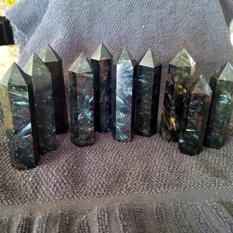 May include: A collection of 12 black and green gemstone points, also known as black tourmaline, arranged on a gray fabric surface. The points are all approximately the same size and shape, and they have a natural, rough texture.