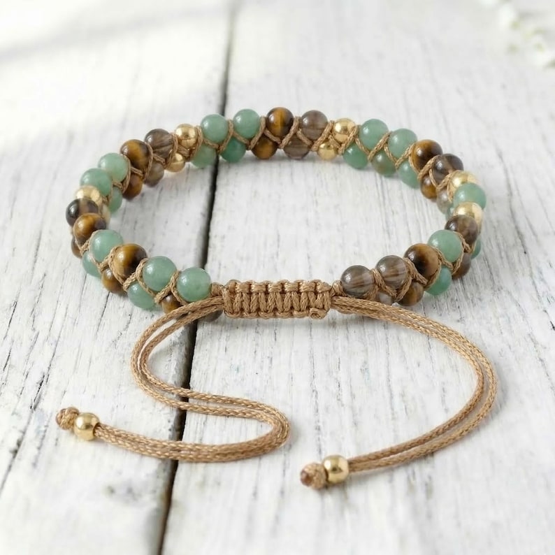 May include: Beaded bracelet featuring green, brown, and gold-colored beads. The bracelet is strung on a tan cord with an adjustable sliding knot closure. The beads are various sizes and shapes, creating a unique and natural look. The bracelet is displayed on a white wooden surface.