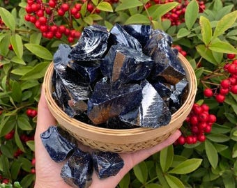 Puede incluir: Una mano sostiene una cesta tejida llena de piedras preciosas azul oscuro y brillantes. Las piedras preciosas tienen una forma rugosa e irregular. Hojas verdes y bayas rojas en el fondo.