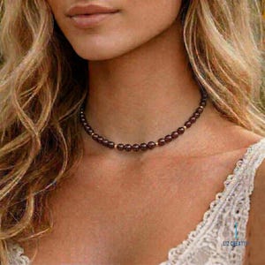 May include: A choker necklace featuring dark red beads and small gold-colored spacers. The necklace is worn around the neck. The background is blurred, with a focus on the necklace and the skin of the person wearing it.