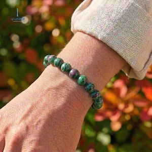 Puede incluir: Un brazalete de cuentas verdes y granates. Las cuentas redondas presentan un patrón moteado. El brazalete se lleva en la muñeca, con una manga de color neutro en el fondo. Es una pieza de joyería.
